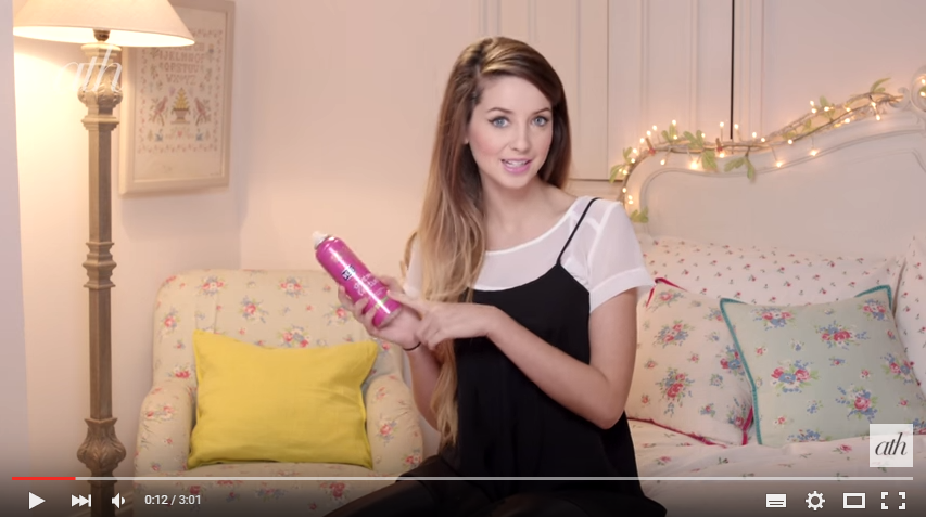 YouTubeのスターZoellaによるフィッシュテールブレードチュートリアルのスクリーンショット。Zoellaがベッドに座って、ピンクの乾いたテクスチャスプレーの缶を持っているところを示しています。