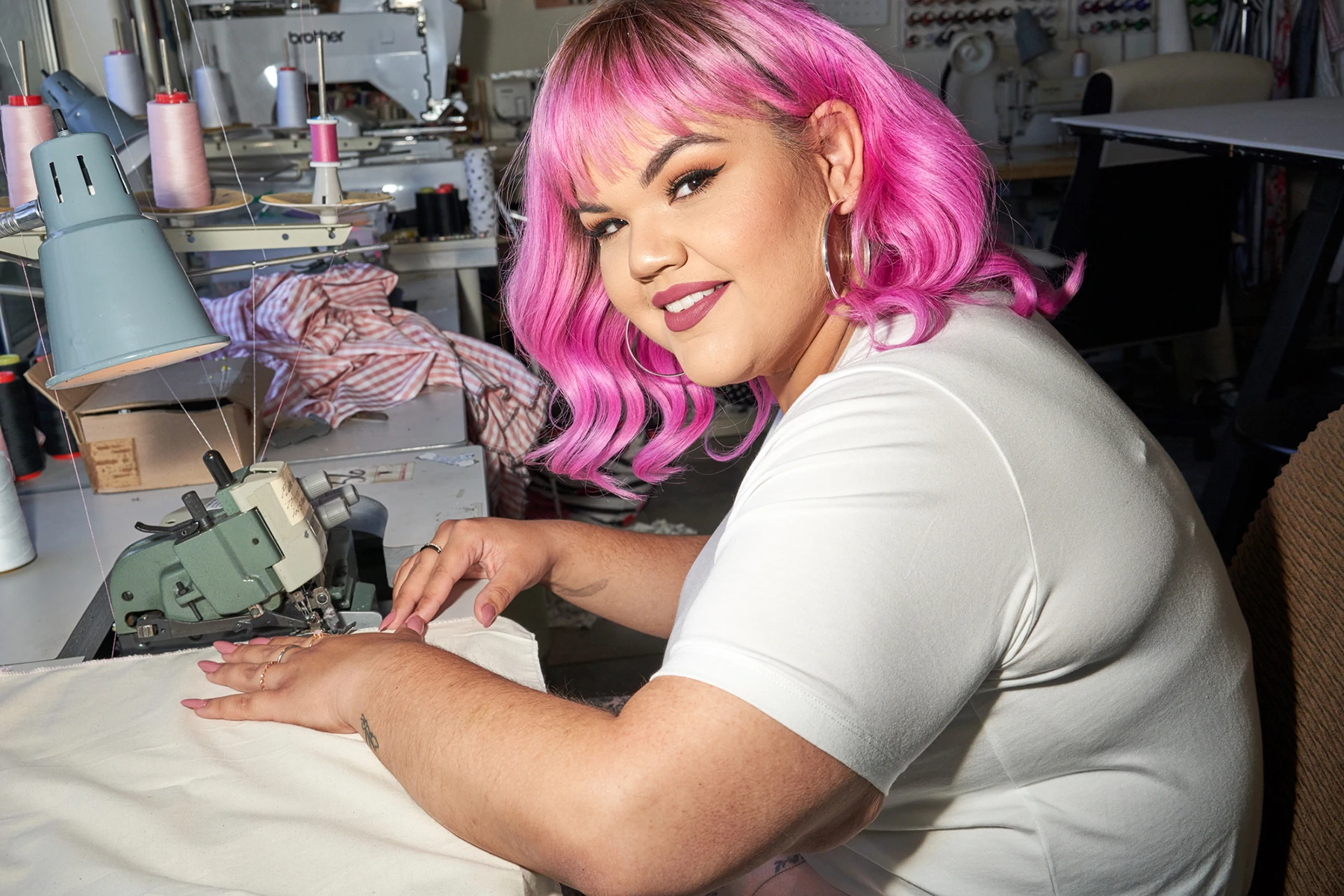 Plus la créatrice de mode Ashley Nell Tipton avec des cheveux roses vibrants, souriant et cousant du tissu.