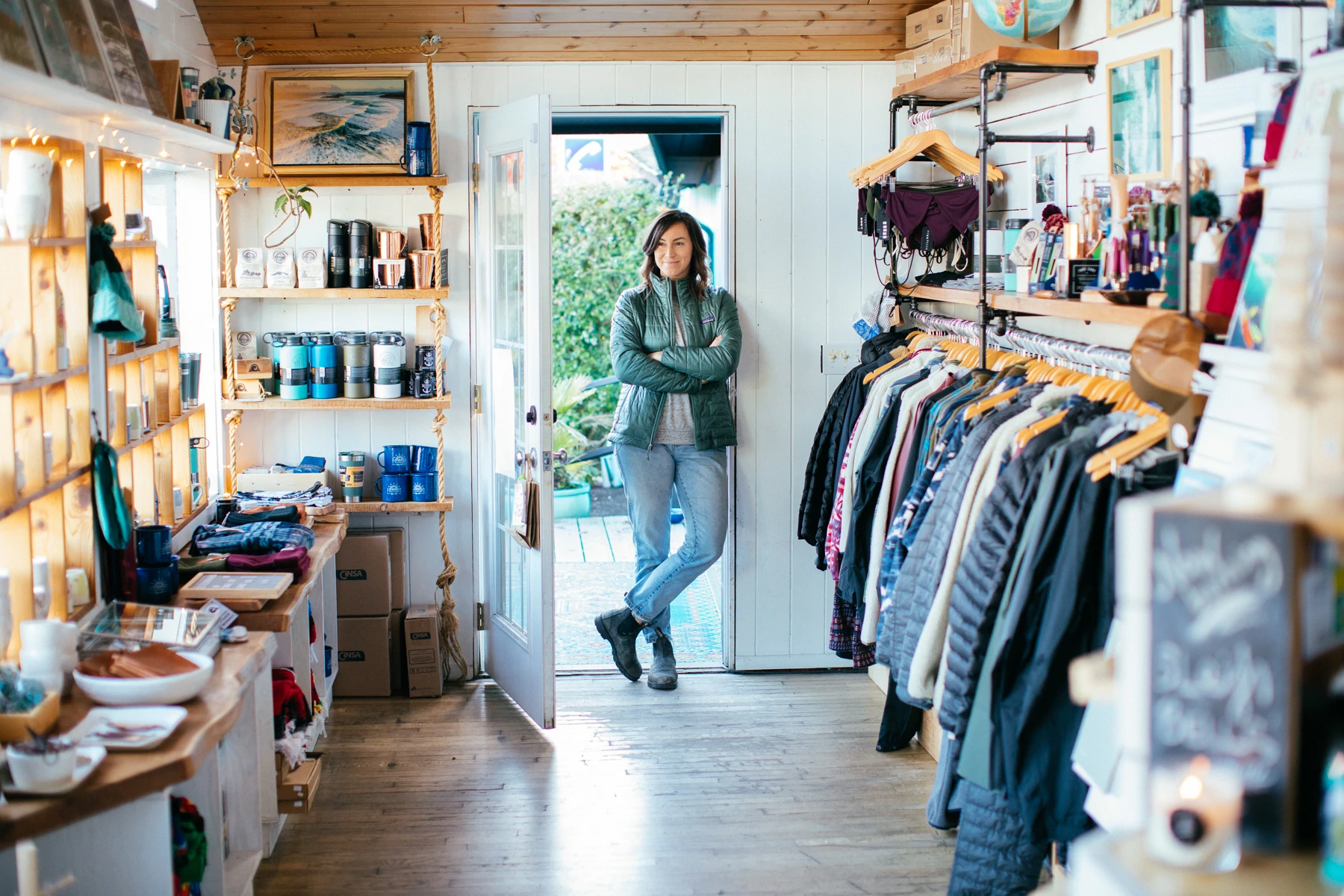 Jen Thorpe, in giacca verde e jeans, è in piedi davanti alla porta del suo negozio, Caravan Beach Shop. Si affaccia sulla collezione di articoli per la casa a sinistra e di vestiti a destra.