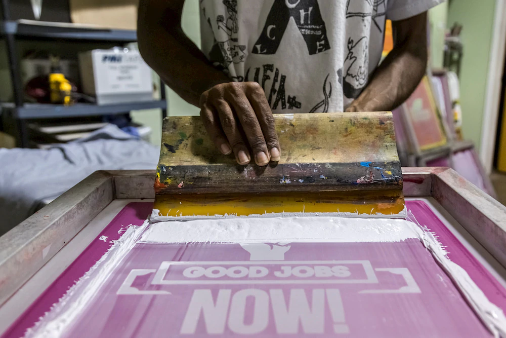 Detaliu al mâinilor unei serigrafi care aplică cerneală pe un ecran pe care scrie „LUCRĂRI BUNE ACUM!”