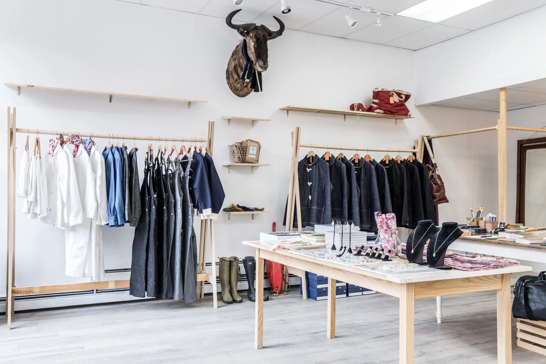 Plan large d'un magasin de détail aéré et minimal avec des luminaires en pin, un décor de taxidermie et des étagères de denim et de vêtements blancs suspendus.