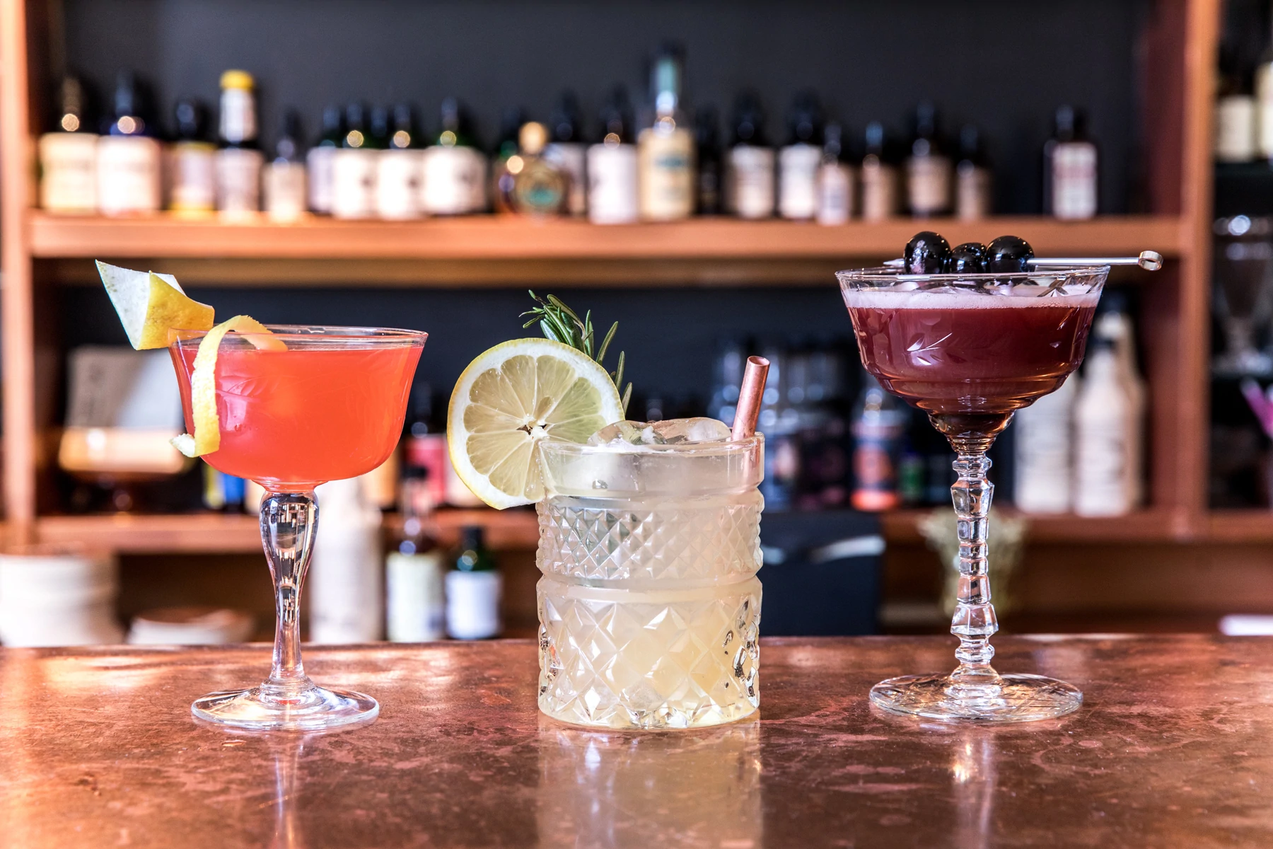 Image de trois cocktails : un orange et garni d'écorces d'agrumes, un jaune pâle et garni de citron et de romarin, un violet et garni de baies. Les boissons sont assises sur un bar recouvert de cuivre avec des bouteilles de spiritueux et d'amers en arrière-plan.