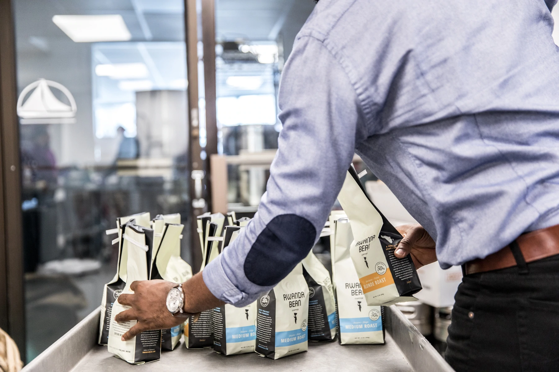 Détail d'un homme empilant des sacs de grains de café sur une table dans une installation de production. Les sacs sont étiquetés "Rwanda Bean".