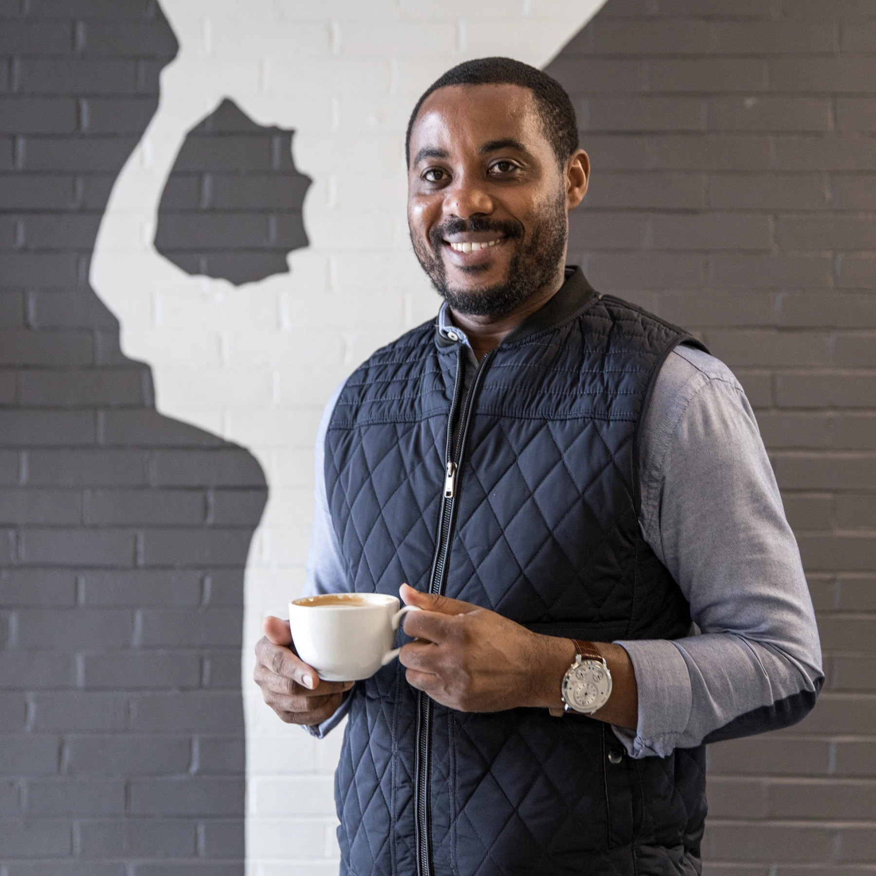 Portrait de Mike Mwenedata, fondateur de Rwanda Bean, tenant une tasse de café et debout devant un mur de briques.