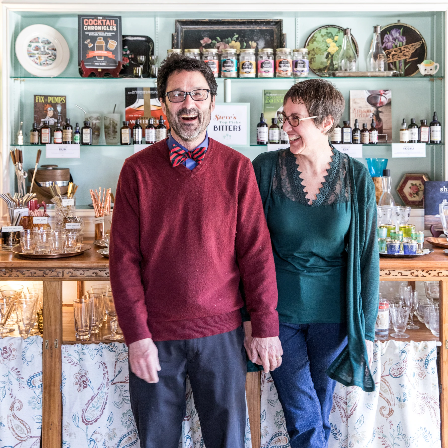 Un homme et une femme se tiennent la main et rient devant un présentoir de verres et d'amers à cocktails.