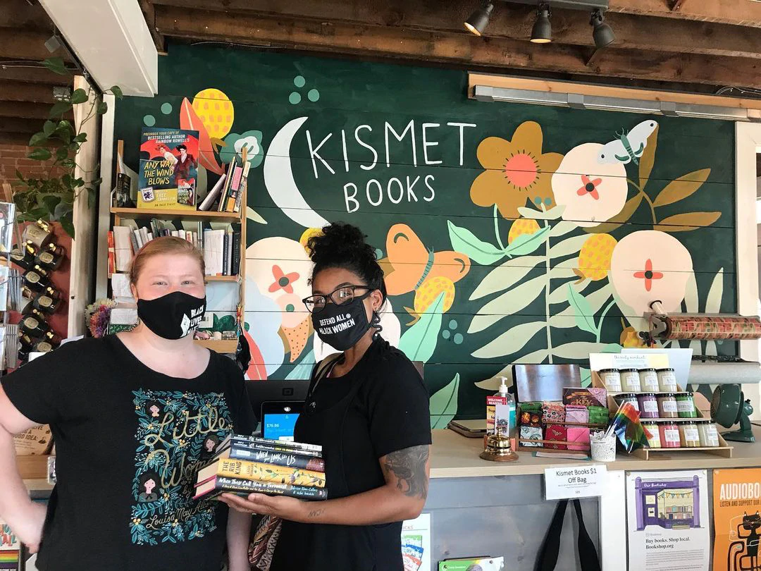 Dominique Lenaye rend visite à une autre libraire dans son magasin. Une fresque murale derrière eux indique "Kismet Books"