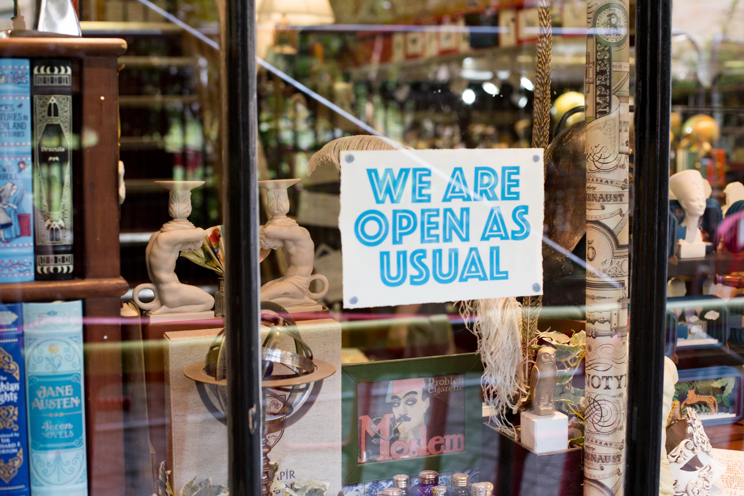 La devanture d'une librairie et de curiosités photographiée de l'extérieur. Un panneau sur la fenêtre indique "nous sommes ouverts comme d'habitude"