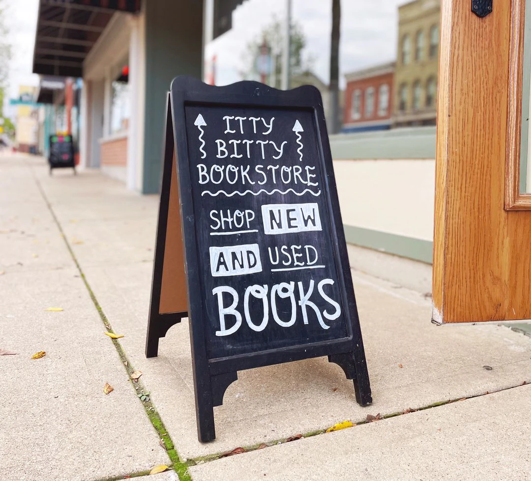 Un panneau sandwich est posé sur un trottoir devant un magasin. Il lit "Itty Bitty Bookstore. Achetez des livres neufs et d'occasion"