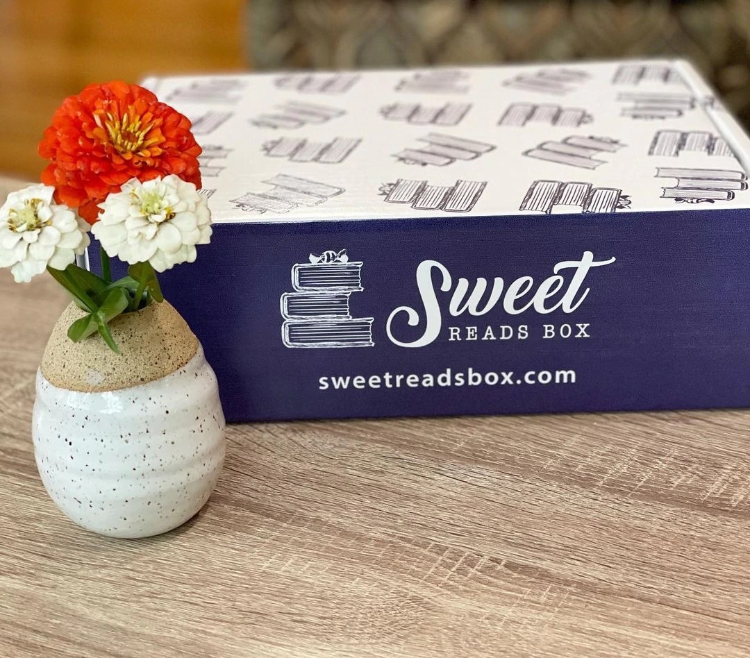 Une boîte d'abonnement imprimée avec le texte "Sweet Reads Box" est posée sur une table à côté d'un vase de fleurs