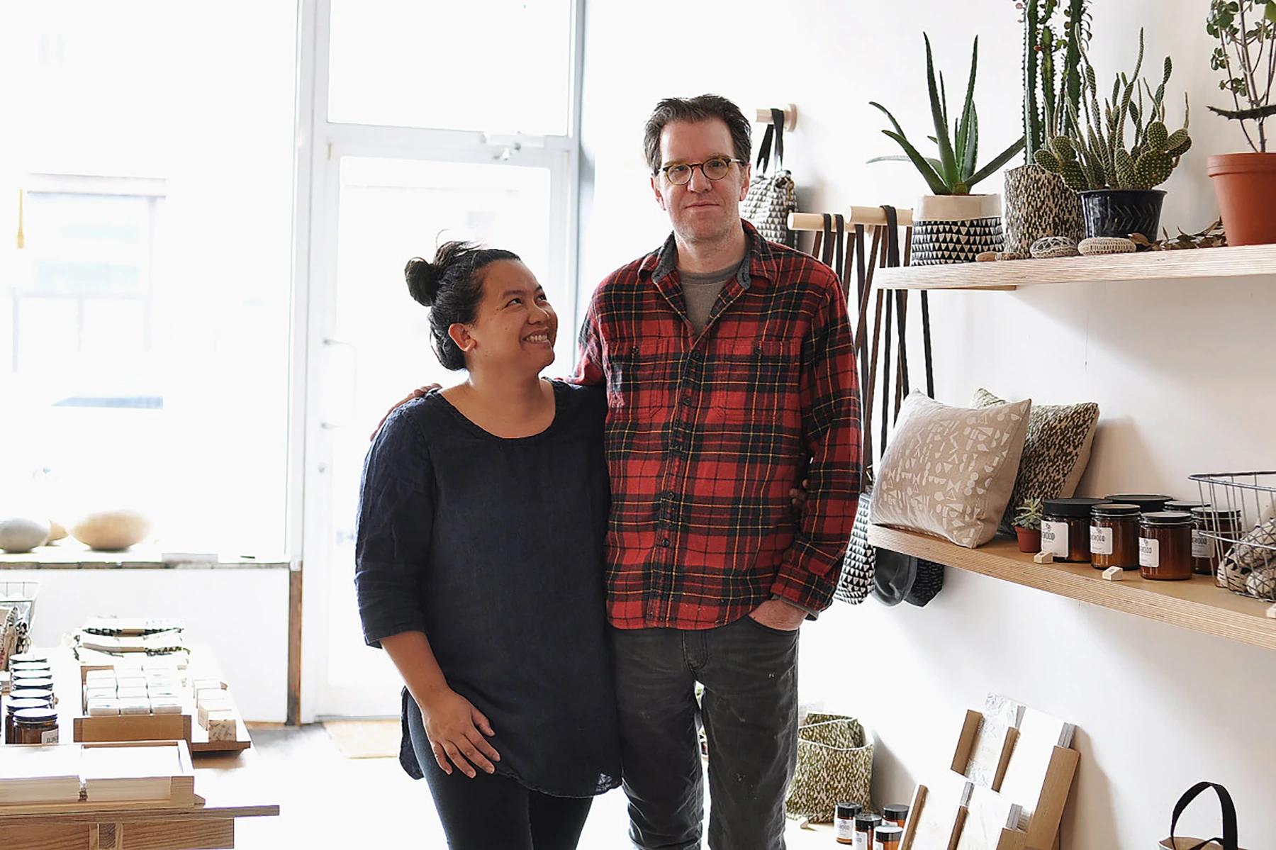 Retrato de Arounna e John, fundadores da Bookhou, em sua loja de varejo. Há produtos artesanais nas prateleiras à direita e Arounna está olhando para um John enquanto olha diretamente para a câmera. Arounna está vestindo um top azul marinho solto de manga comprida e John está vestindo uma camisa xadrez vermelha de madeira.