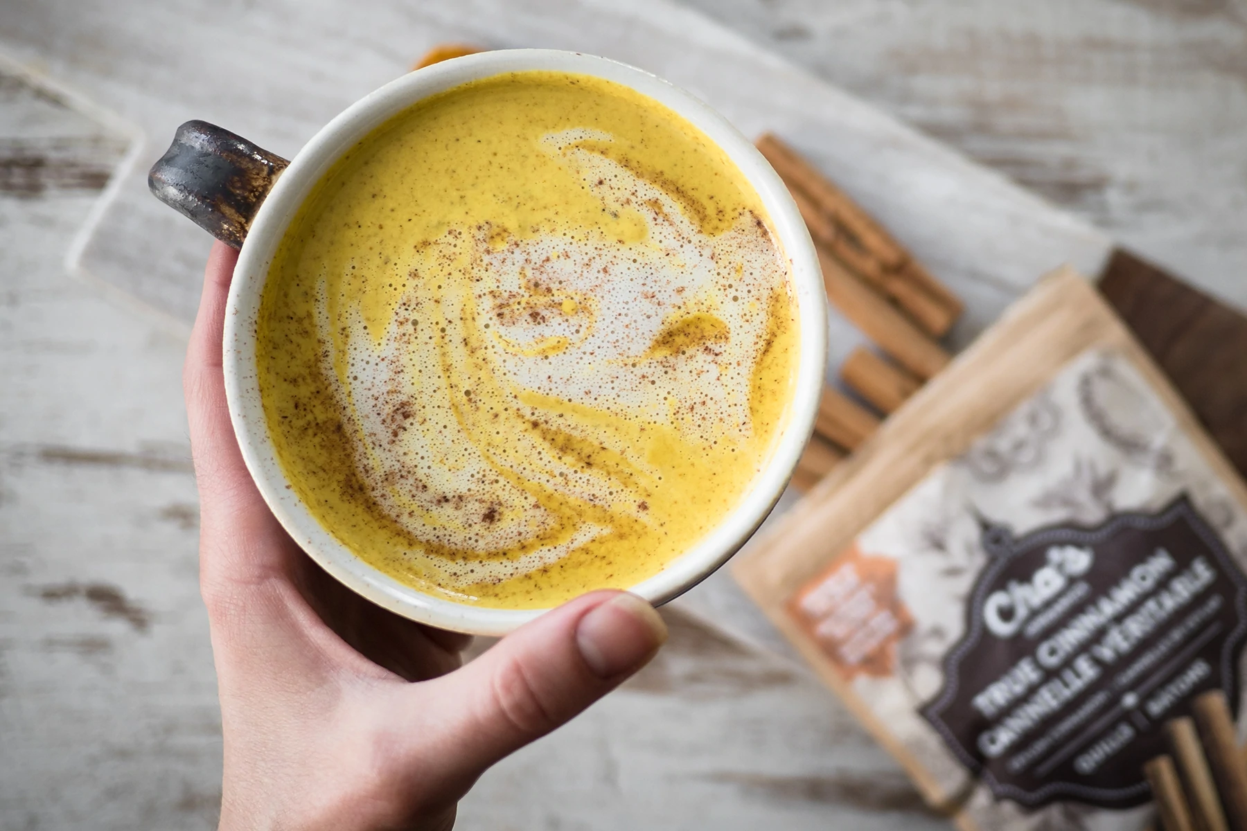 Fotografia aérea da mão de uma mulher caucasiana segurando uma xícara de chai latte da Cha's Organic, sobre uma superfície de madeira. Ao fundo, na mesa sob sua mão, está o saquinho de chá Cha's Organics.