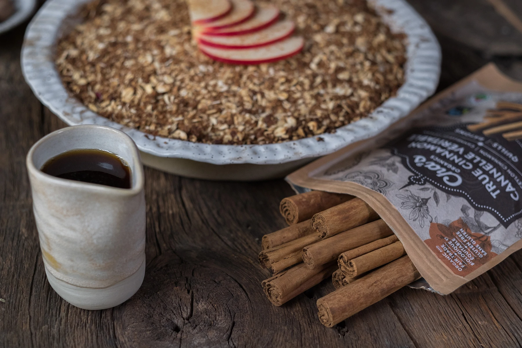 Fotografia dos paus de canela da Cha's Organics abertos com alguns paus derramando sobre uma superfície de madeira. Ao lado dela há um pequeno recipiente de xarope de bordo e uma tigela de cereal quente ao fundo, um pouco fora de foco.