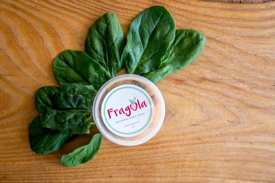 Un petit récipient portant l'inscription "Fragola bio baby food" est posé sur une surface en bois entourée de feuilles d'épinards