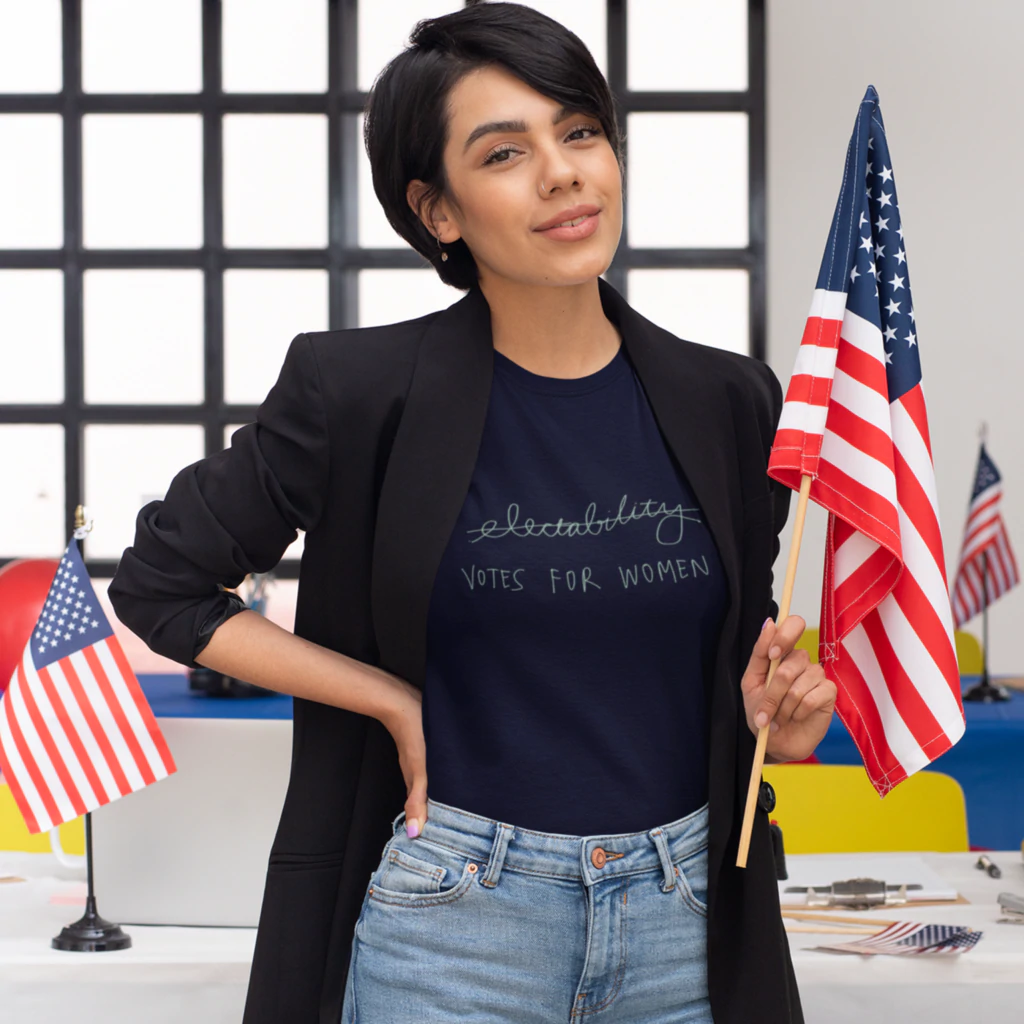 Wanita berdiri di depan meja dengan bendera Amerika. T-shirtnya bertuliskan "suara untuk wanita"