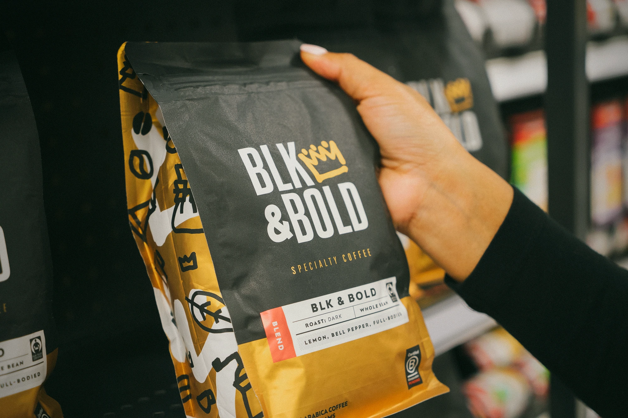 Paket kopi BLK & Bold dipegang dengan tangan di beberapa rak ritel.