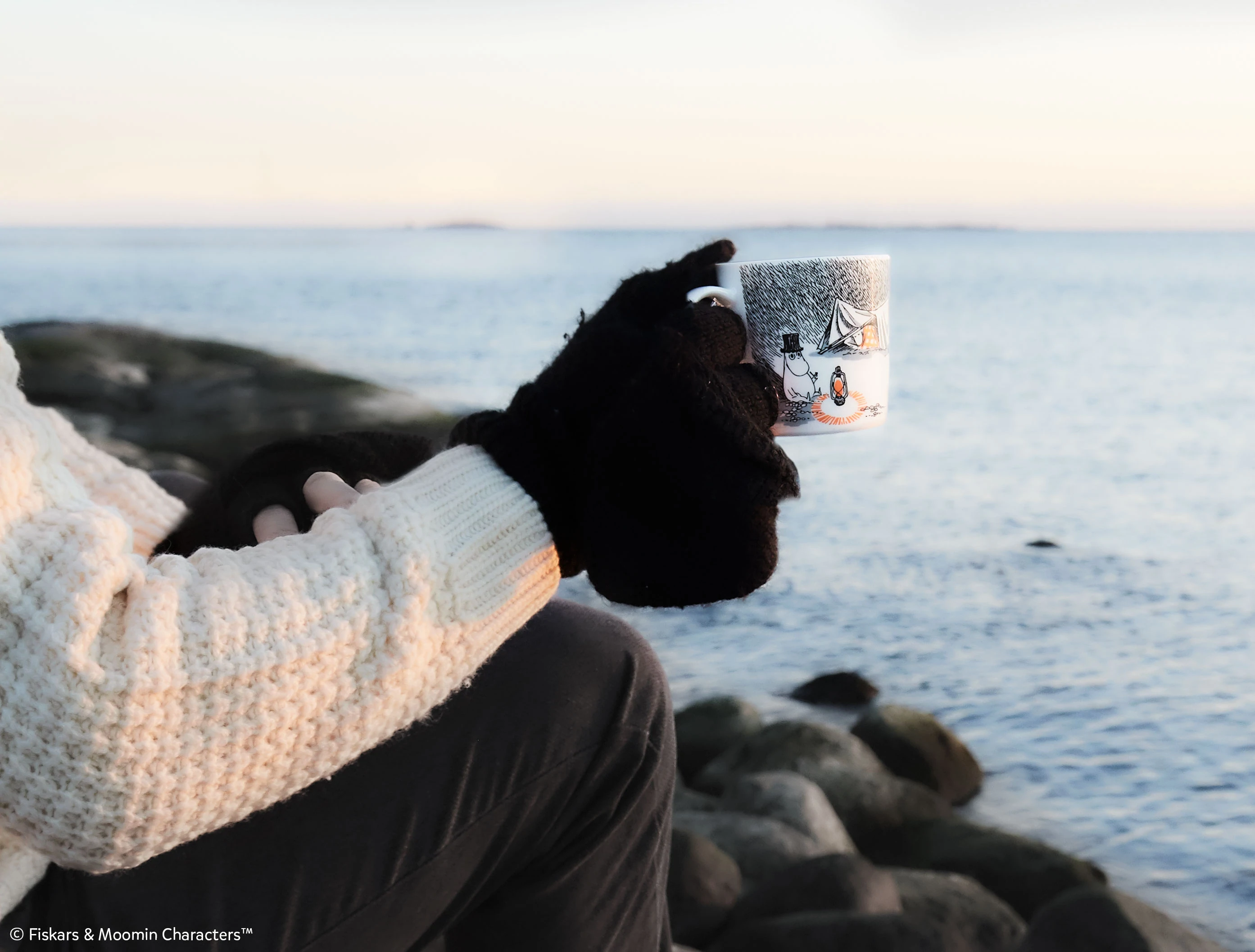 Cangkir Moomin dipegang oleh model tangan, berlatar belakang laut.