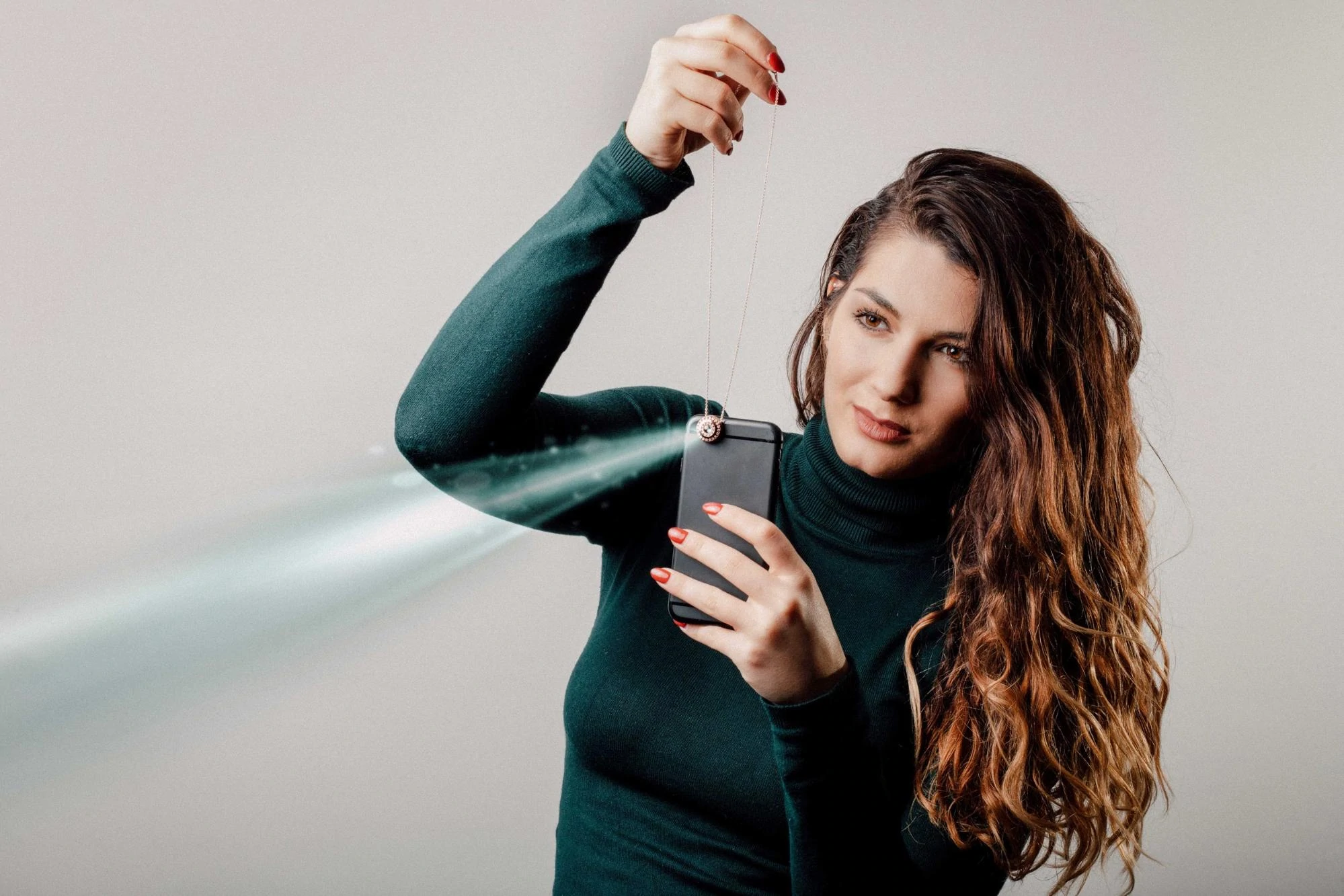 Una modella con un maglione verde tiene in mano un medaglione di gioielli Revival mentre fa brillare una luce dal suo cellulare per proiettare un'immagine dalla gemma del ciondolo.