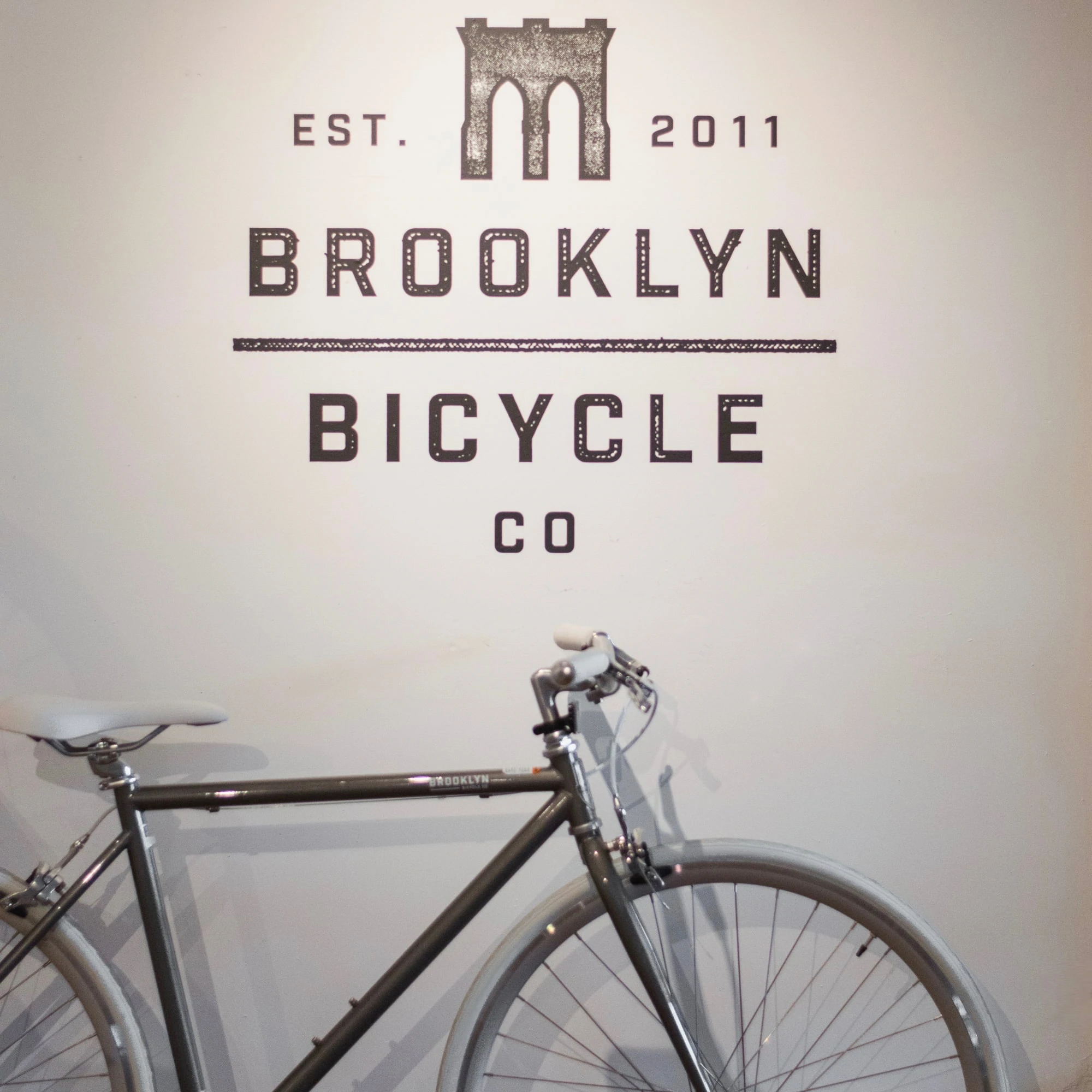 Una bicicletta grigia di Brooklyn Bicycle Co. nel loro showroom contro un muro con il loro logo.