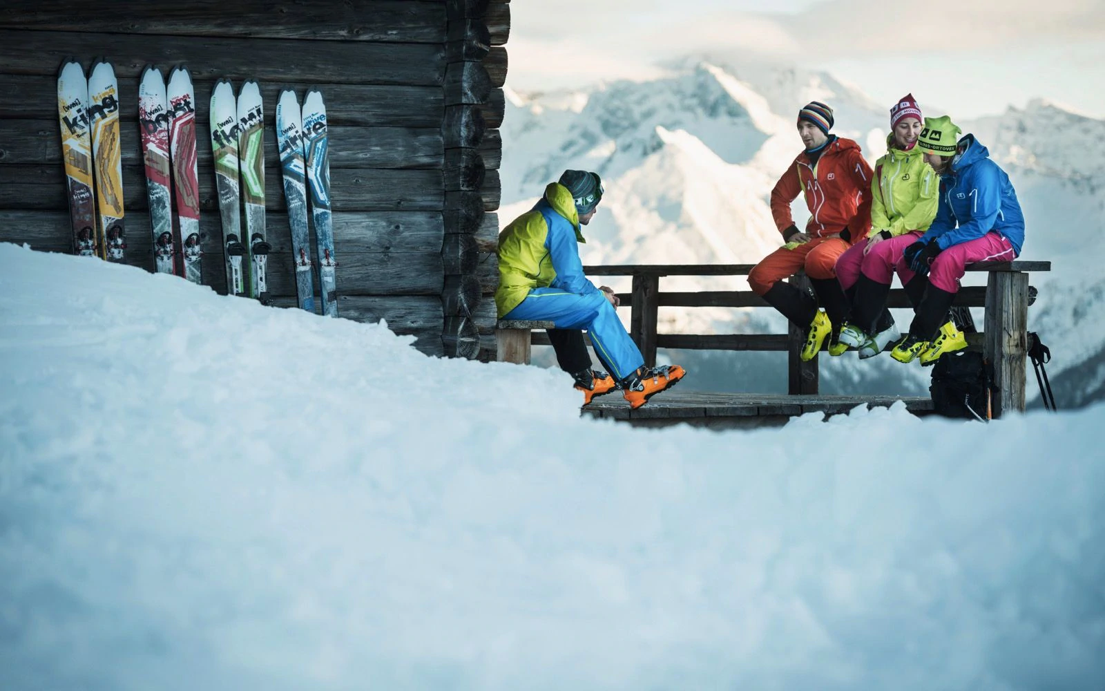 Un gruppo di quattro sciatori seduti fuori da uno chalet che ha paia di sci appoggiati sul lato della parete.