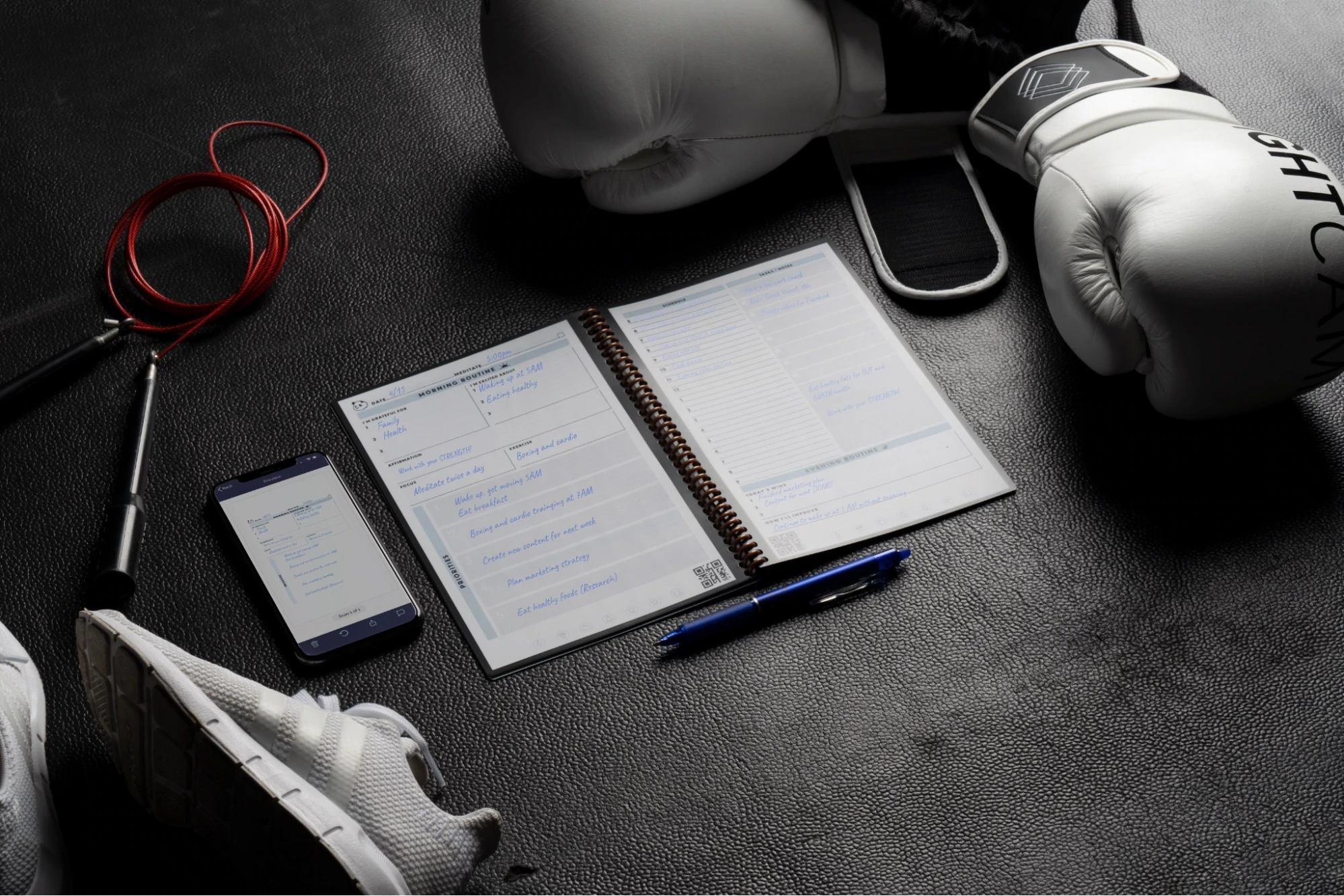 Ein Rocketbook-Notizbuch zusammen mit einem Smartphone, das seine App anzeigt, im Hintergrund Trainingsausrüstung wie Boxhandschuhe und Laufschuhe.