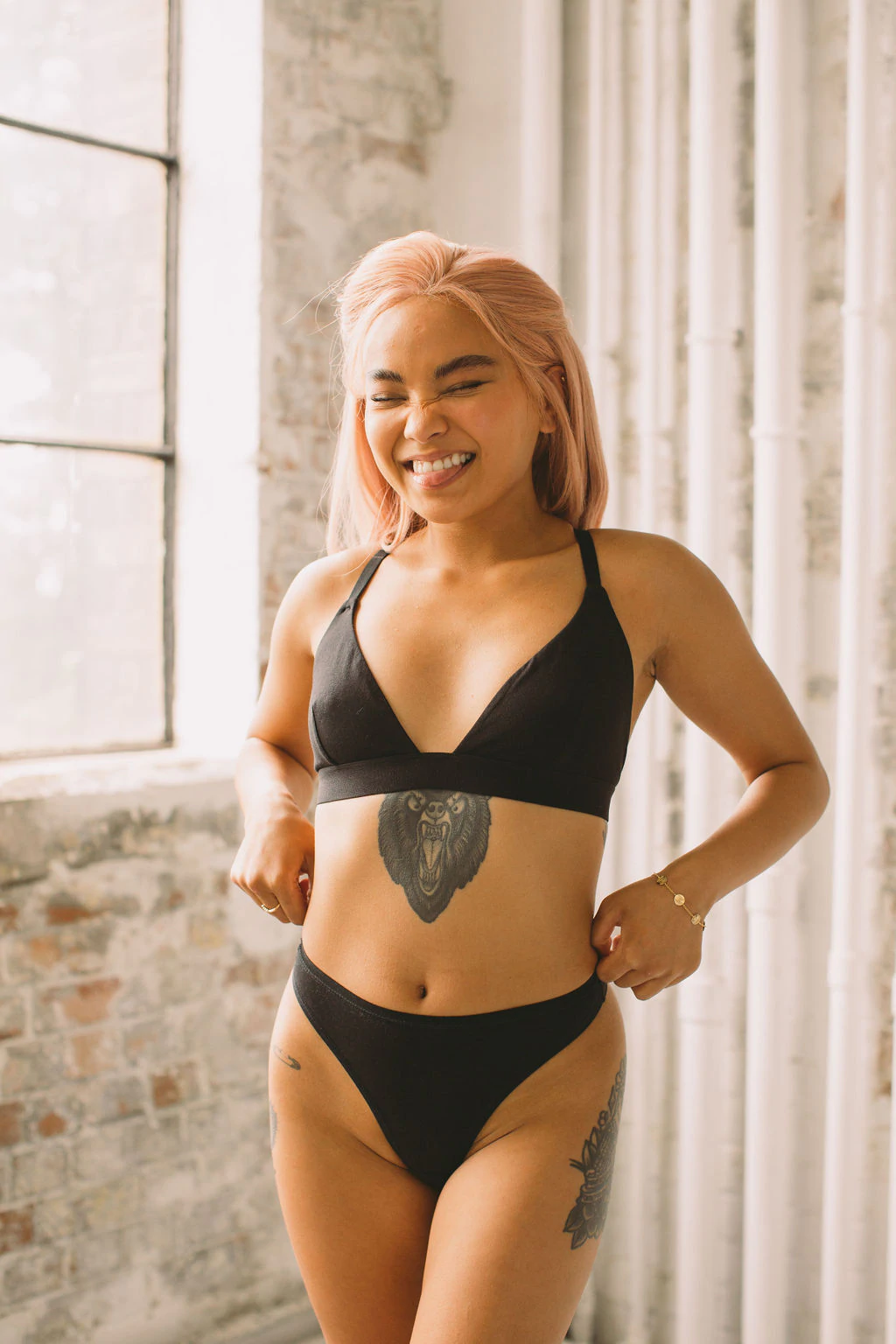 Seorang model berpose di depan jendela mengenakan bralette segitiga hitam dan thong set oleh Pantee