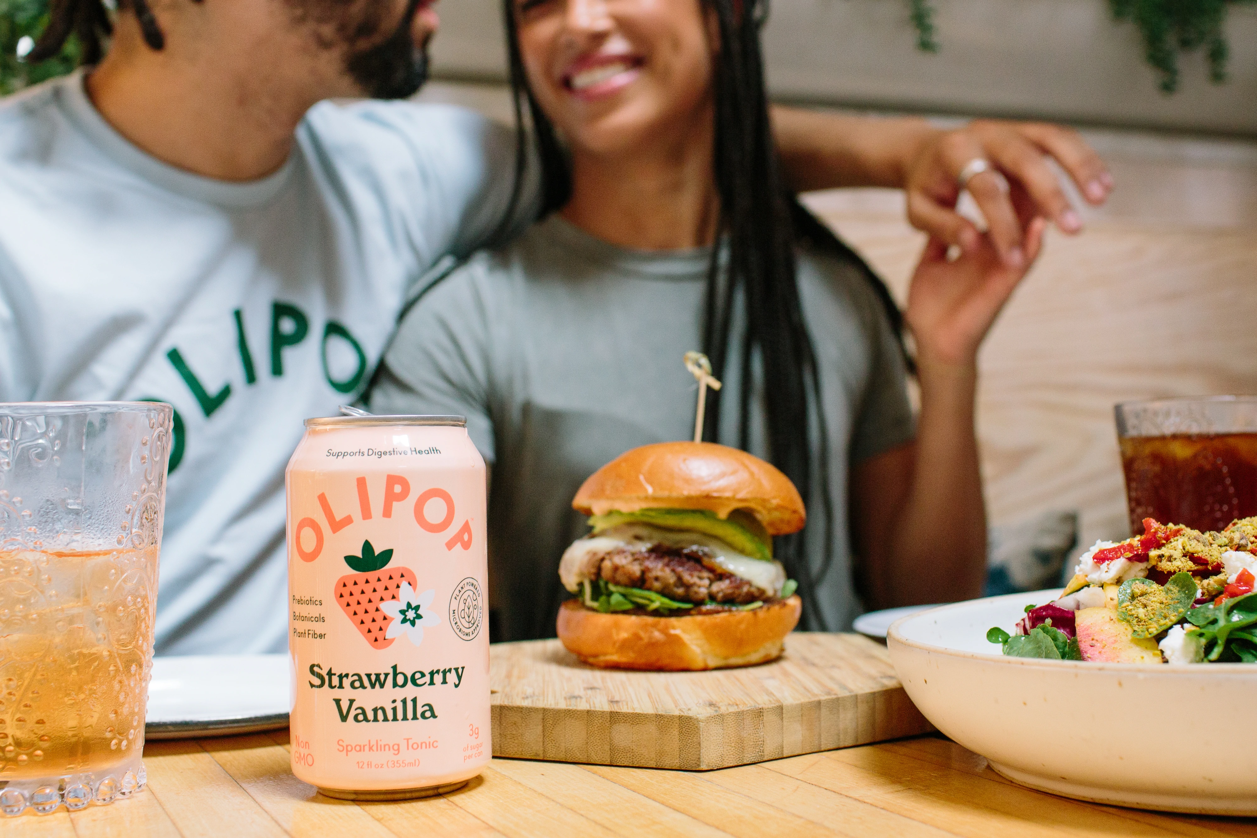 Önlerinde hamburger ve bir kutu Olipop olan bir restoranda otururken kucaklaşan bir çift.